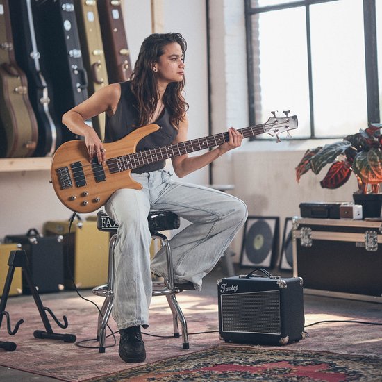 Een vrouw met donker haar speelt op een houten basgitaar, zittend op een krukje. Naast haar staat een kleine Fazley versterker.