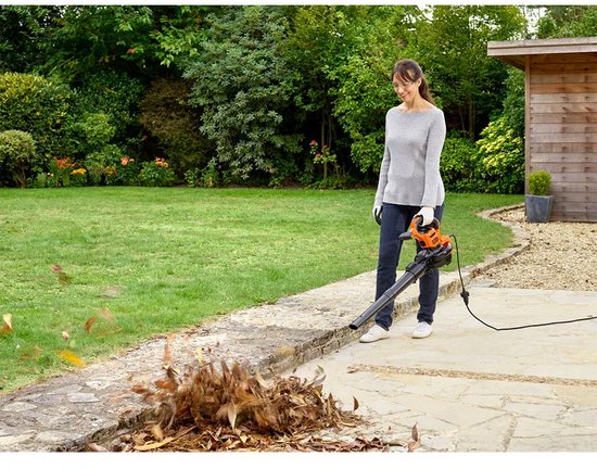 Vrouw gebruikt oranje bladblazer, blaast bladeren van een stenen pad in de tuin.