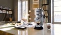 Zilverkleurig De'Longhi espressoapparaat op een glanzende tafel in een woonkamer, met een glaasje espresso en een glas cappuccino.