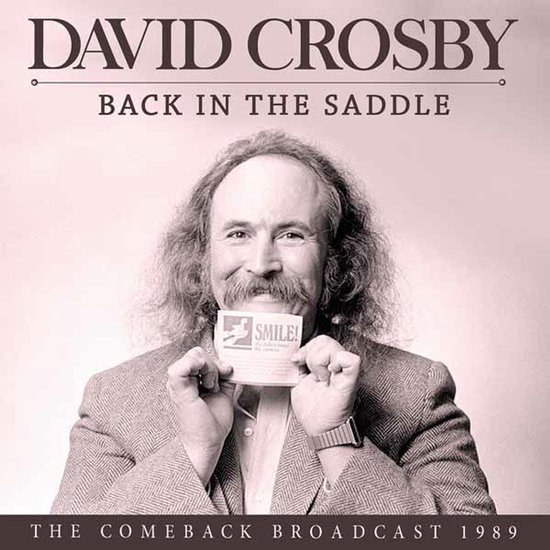 David Crosby met snor en krullend haar, met grijze blazer, houdt een kaartje omhoog met de tekst "SMILE!". Tekst: "DAVID CROSBY BACK IN THE SADDLE THE COMEBACK BROADCAST 1989"