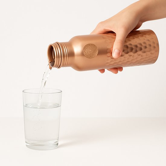 Een hand giet water uit een koperen fles met hamerslag textuur in een glas.