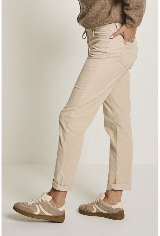 Een beige corduroy broek met koordjes en omgeslagen pijpen, gedragen door een model met bruine trui en sneakers.