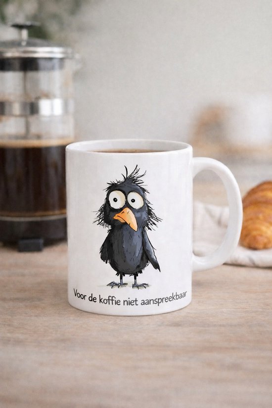 Witte mok met een tekening van een zwart vogeltje met warrig haar en grote ogen. Tekst: "Voor de koffie niet aanspreekbaar".