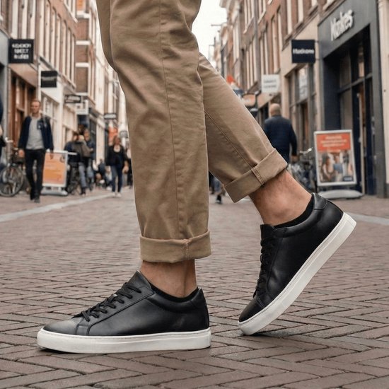 Man in bruine broek en zwarte sneakers met witte zolen, staand op een straat.