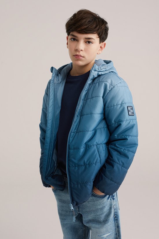 Jongen in een blauwe gewatteerde jas met een badge op de mouw, spijkerbroek.