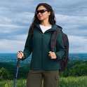 Vrouw met groenblauwe softshell jas, wandelstokken en rugzak op een heuveltop.