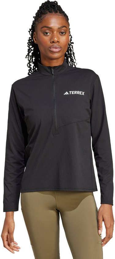 Model draagt een zwart sportshirt met halve rits en beige leggings, focus op Terrex-logo.