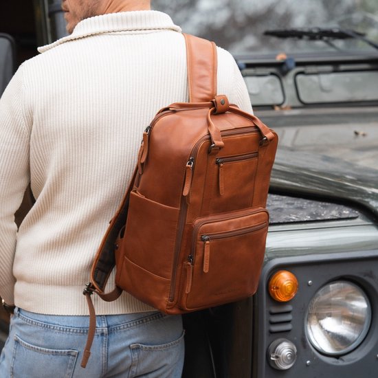 Man draagt een cognac leren rugzak, staand naast een donkere terreinwagen.