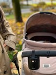Zwarte ZAZU Baby Rocker bevestigd aan de duwstang van een lichtbruine kinderwagen, naast een persoon.