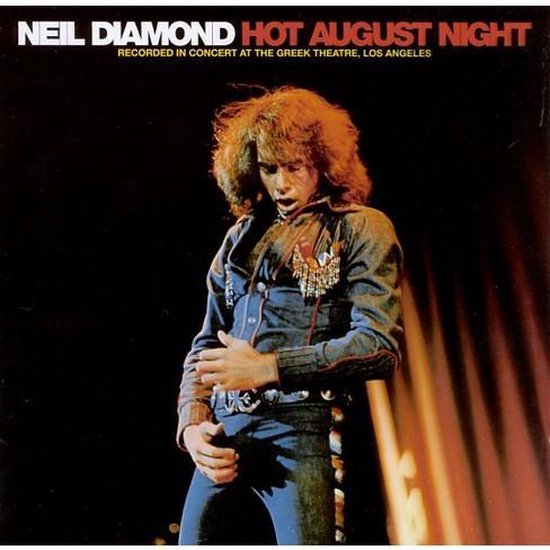 Neil Diamond met lang bruin haar, in een spijkerjasje met lovertjes en spijkerbroek, op een podium.