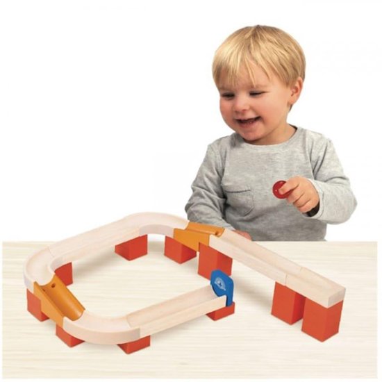 Een jongen met blond haar kijkt naar een houten knikkerbaan met oranje blokken en een rode bal.
