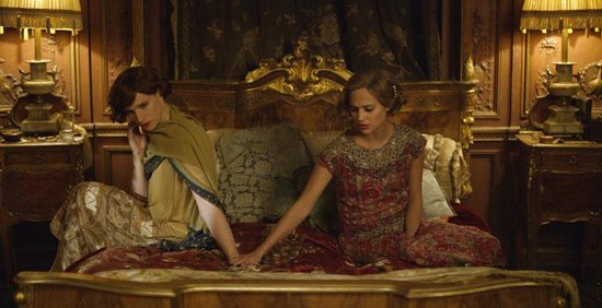 Twee vrouwen zitten op een bed. De ene vrouw is gekleed in een gele sjaal, de andere in een rood-geprint patroon.
