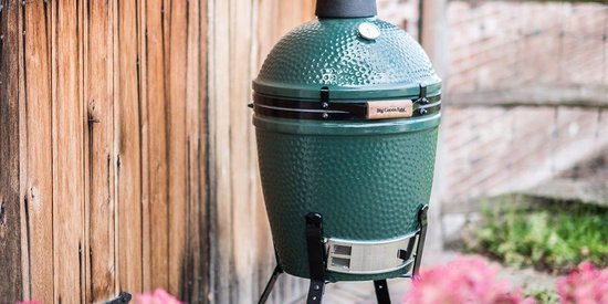 Donkergroene Big Green Egg barbecue met houten handvatten op een standaard, naast een houten schutting.