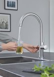 Chroom GROHE kraan schenkt heet water in een glas met theezakje, met de hand op de hendel.