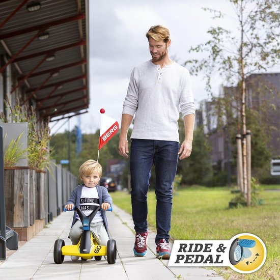 Man loopt naast een kind dat op een gele loopauto met BERG-vlag rijdt, buiten bij een modern gebouw.