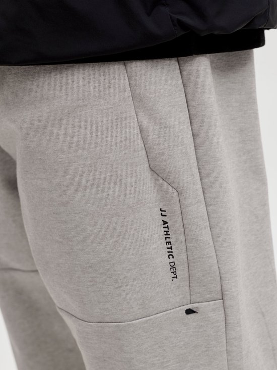 Close-up van een grijze joggingbroek met een ritsvak en 'JJ Athletic Dept.' opdruk.