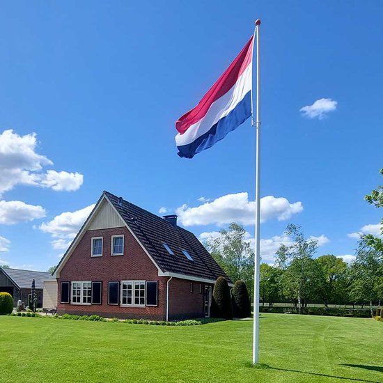 Nederlandse vlag op een witte mast, naast een bakstenen huis met schuur, op een groen gazon onder een blauwe lucht.