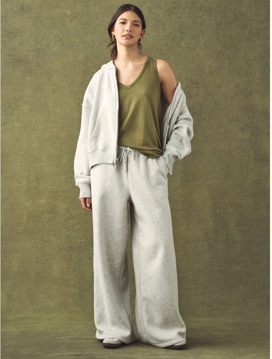 Een vrouw met bruin haar draagt een groen mouwloos topje, een grijze oversized hoodie, en een wijde grijze joggingbroek.