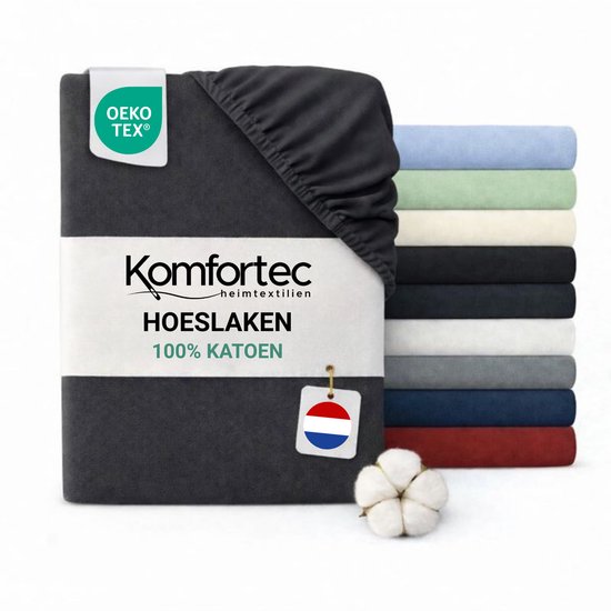 Donkergrijs katoenen hoeslaken van Komfortec, met OEKO-TEX label. Op de achtergrond stapels hoeslakens in verschillende kleuren.