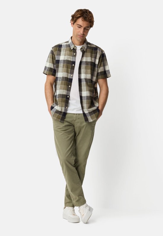 Man in geruit groen overhemd met korte mouwen, wit T-shirt en olijfgroene broek.