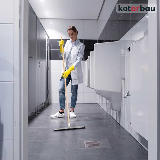 Een vrouw met gele handschoenen gebruikt een vloertrekker met houten steel in een toiletruimte.