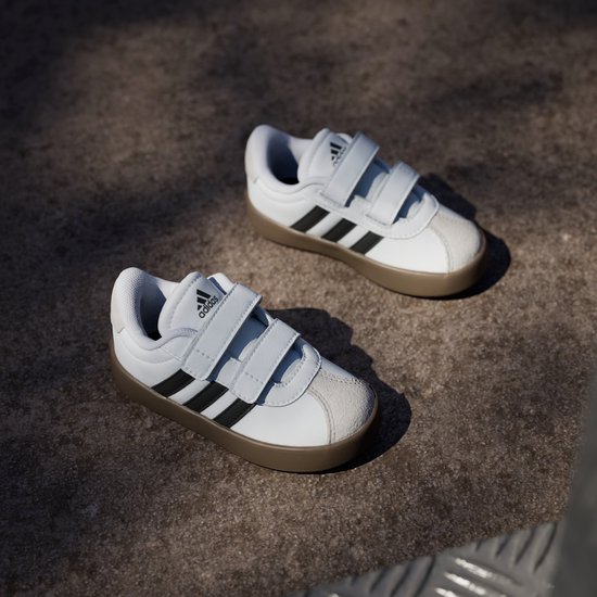 Twee witte sneakers met zwarte strepen, klittenbandsluiting en bruine zool, schuin van bovenaf.
