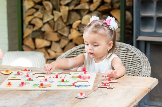 Klein meisje met strikjes in haar haar, zit aan een tafel en legt een houten boerderijpuzzel.