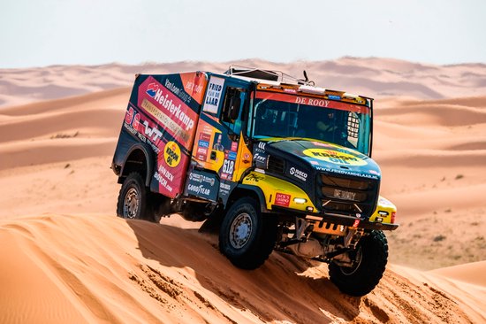 Een grote gele en zwarte rallytruck met sponsorslogo's, waaronder KROON OIL, rijdend door woestijnzand.