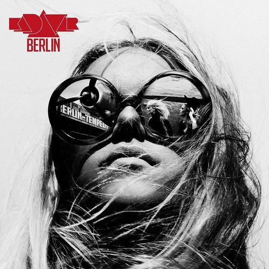 Gros plan en noir et blanc d'une femme aux cheveux flottants et grosses lunettes de soleil, reflétant un avion. Logo 'BERLIN'.