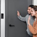 Man en vrouw zwaaien bij de deur, terwijl de zwarte Aqara deurbel aan de muur hangt.