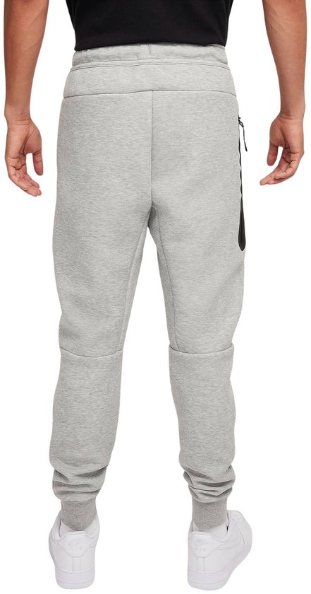 Man draagt lichtgrijze joggingbroek, achterkant met geribde manchetten en zwarte rits op rechterpijp.