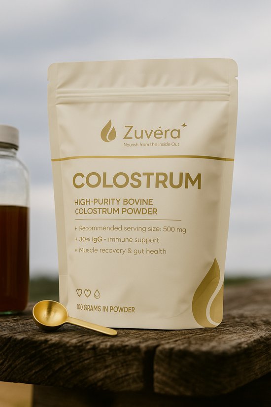 Beige zak "Zuvéra Colostrum" op een houten tafel, naast een glazen fles. Een gouden lepel met poeder ligt ervoor.