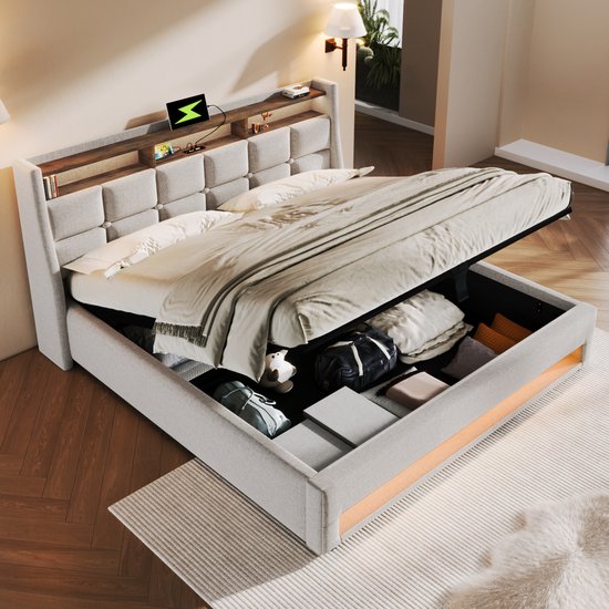 Opbergbed met openklapbare lattenbodem, gevuld met kussens en tassen. Beige gestoffeerd hoofdbord met opbergvakken.