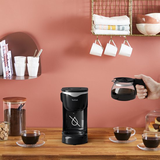 Une main tient une verseuse à café en verre pleine au-dessus d'une cafetière Tefal noire. Deux tasses de café sont prêtes.