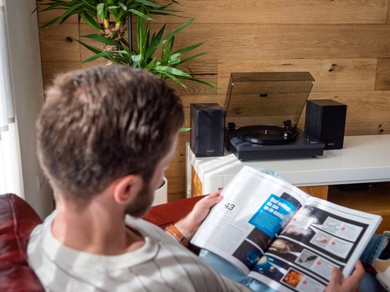 Mann luistert naar muziek op een platenspeler met twee zwarte speakers, in een woonkamer.