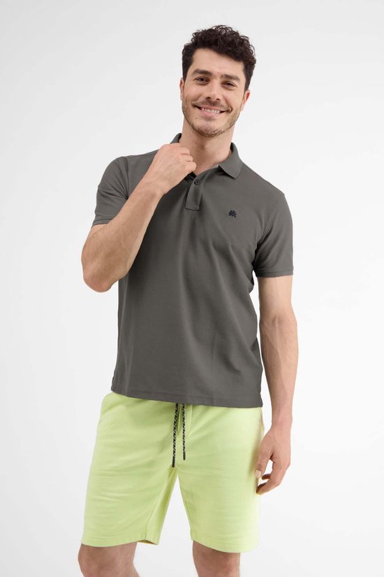 Man in donkergrijs poloshirt en lichtgroene korte broek, glimlacht en raakt kraag aan.