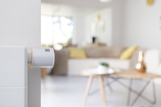 Een witte Netatmo slimme radiatorthermostaat met oranje accent, gemonteerd op een radiator in een woonkamer.