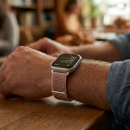 Een pols met een smartwatch en een roze stoffen horlogebandje. De persoon zit aan een houten tafel.