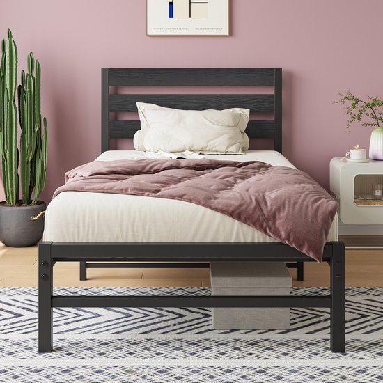 Lit en métal noir préparé avec tête de lit en bois, avec un oreiller blanc, une couverture rose et un magazine, sur un tapis graphique.