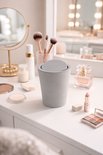 Grijze miniprullenbak met deksel op een make-uptafel, met cosmetica en parfum.