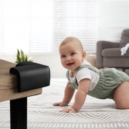 Een lachende baby kruipt op een vloerkleed naar een zwarte hoekbeschermer op een houten tafel.