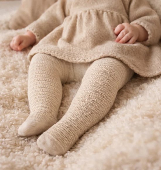 Baby draagt beige gebreide maillot met textuur onder een bijpassende jurk op een zachte ondergrond.