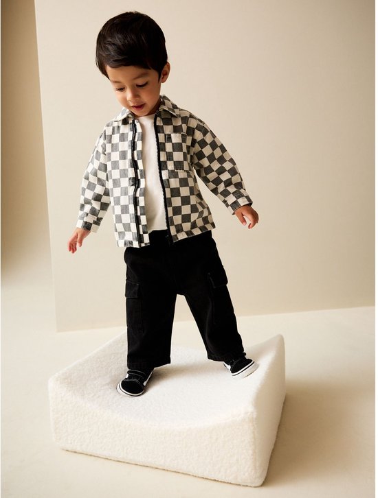 Jongen in een grijs-wit overshirt met rits en zwarte broek, staand op een witte verhoging.