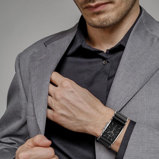 Man met grijs pak en zwart overhemd draagt een rechthoekig zwart keramisch horloge met zilverkleurige wijzerplaat.