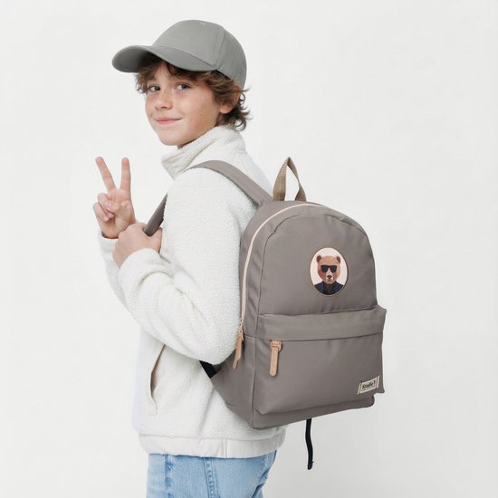 Jongen met grijze pet en witte jas draagt een grijze rugzak met beer-patch.