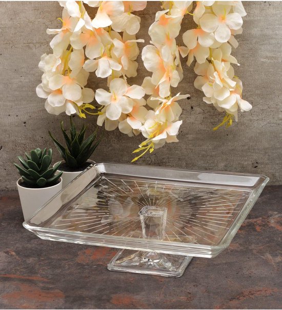 Vierkante glazen taartplateau met decoratieve lijnen en een verhoogde voet. Twee plantjes staan ernaast.
