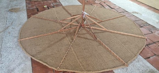 Een ronde parasol van kokosvezel, met het houten frame zichtbaar aan de onderkant, ligt plat op de grond.