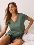 Vrouw op bed, lachend, in een groen V-hals T-shirt en een short met bladerenprint.