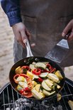 Een persoon met schort bakt groenten, waaronder courgette en paprika, in een Skottsberg pan boven een barbecue.
