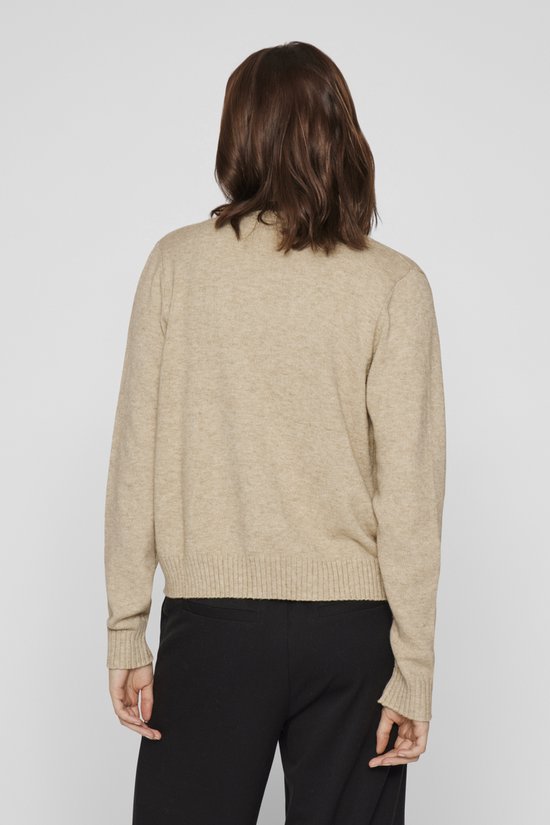 Lichtbeige gebreid vest met lange mouwen, gedragen over een wit shirt en zwarte broek.